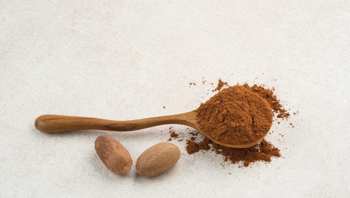 4 épices Poudre de quatre épices dans une cuillère en bois rustique avec noix de muscade entière, sur fond textile blanc texturé, photographie culinaire minimaliste