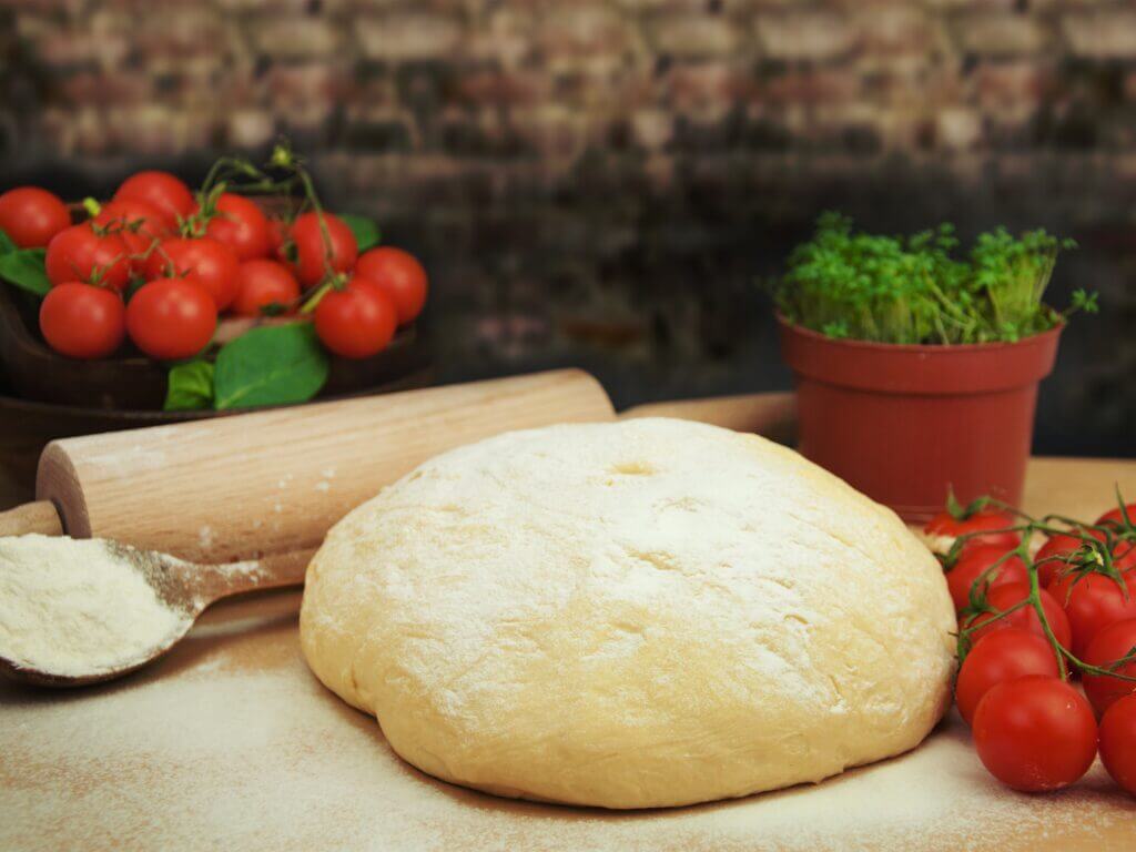 Pizzateig Rezept: Fertig gekneteter Teig mit Mehl, Nudelholz und frischen Kirschtomaten